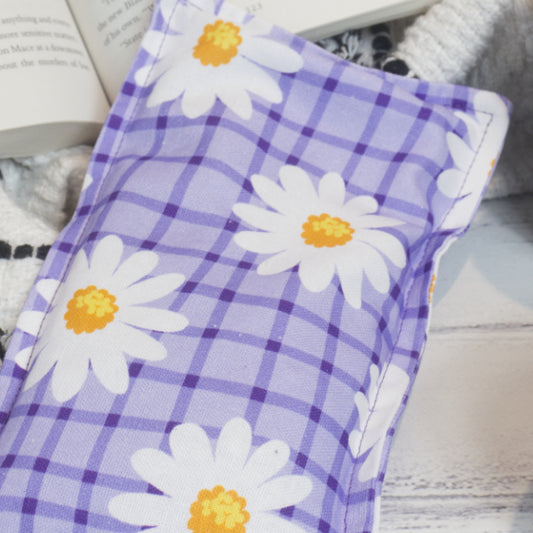 A close up photo of the Purple Daisy Mini Heat pack showing the fabric design of a Purple checkered background with white daisies with yellow centres scattered over. The edge of the heat pack rests against a book and black and white blanket.