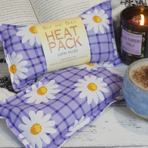 Two Purple Daisies Mini Heat Pack by Soy and Shea rests near a ceramic cup, a burning candle, and an open book—creating an inviting cosy scene. The Mini Heat packs feature a purple checkered pattern with white daisies featuring yellow centers scattered across it.