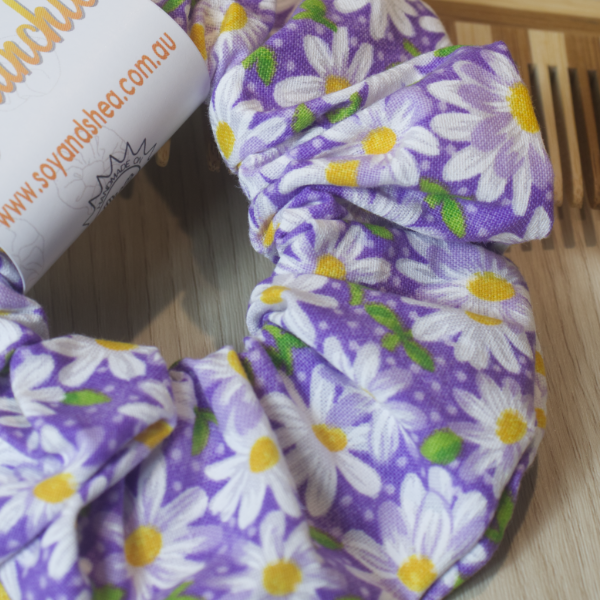 A close up of the fabric design on the Purple Daisy scrunchie showing a purple fabric with white daisies with yellow centers and green leaves scatter throughout.