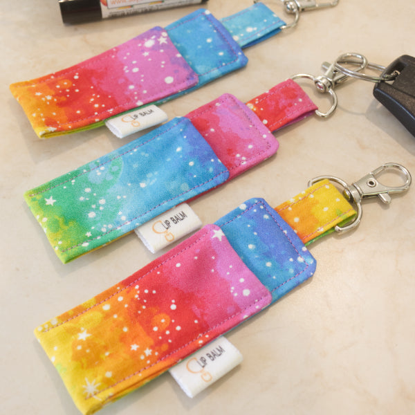 A selection of three clip balms showing variations in fabric design, lie on a wooden side with the middle Clip Balm attached to a car key. A lip balm lays near-by along with some greenery. The Clip balms have a bright layered rainbow design with white speckles. Each shows a slight variation in pattern design
