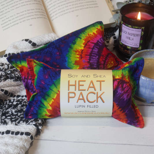 Two Mini Heat packs are stacked upon each other, one with a belly band label reading HEAT PACK LUPIN FIlled. These are resting on a black and white blanket with open book, candle and coffee cup evoking a cosy feel. The Heat packs The heat packs show a tie dies design of concentric circles in rich rainbow colours