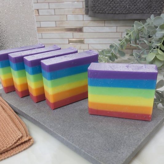 Rainbow striped soap bars on a gray surface with a tiled wall and plant in the background. The soaps feature six even layers of rainbow colour starting with red at the bottom and finishing with purple