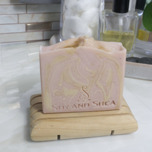 A pale pink soap bar with cream swirls stands on a wooden soap rack on a bathroom countertop with a blurred background