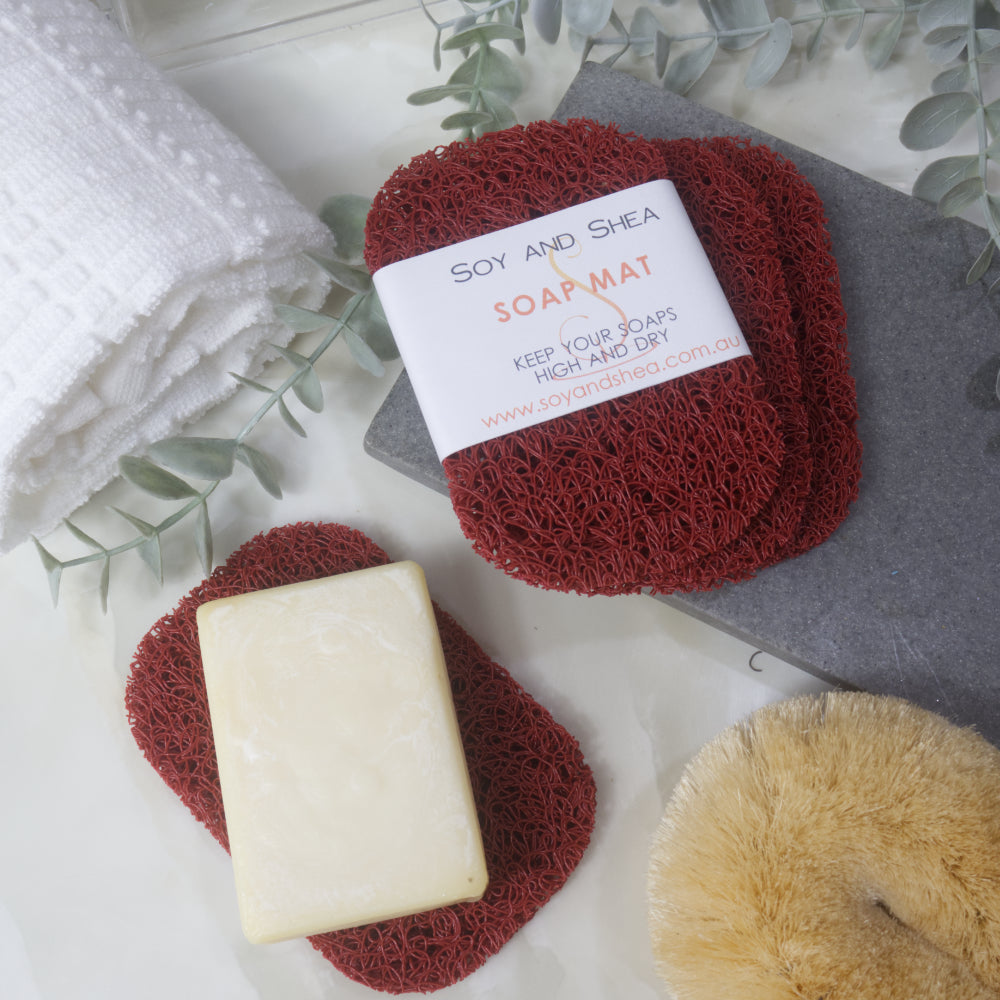 A flatlay image showing a collection of Red Soap mats, displayed on a bathroom countertop along with a white towel, body brush and greenery. Three soap mats are stacked upon each other in a fan shape on a grey stone tray with the top one featuring a product label. A fourth mat lies to the bottom of the image with a cream soap lying on it.