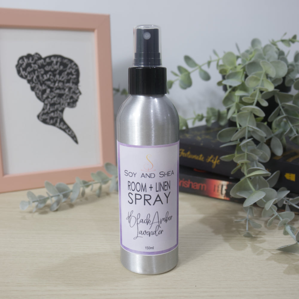 An aluminium bottle with black spray cap stands on a wooden surface surround by a pile of books with a plant sitting on top of them and a picture frame to the left. The bottle features a decorative product label identifying it as Room + Linen Spray, scented in Black Amber Lavender