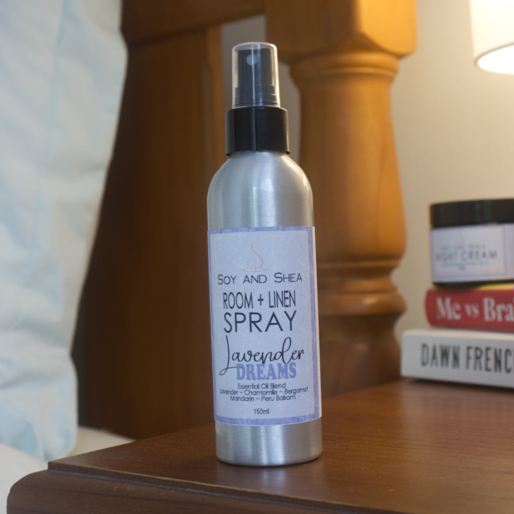 A silver metal bottle with black atomiser lid stands on a wooden beside table along side a stack of books and glowing lamp. The bottle has a white label with purple border and product details.