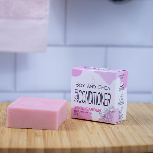 On a wooden tray sits a pink bar of conditioner next to a white box with a pink abstract pattern reading SOLID CONDITIONER against a blurred tiled background.