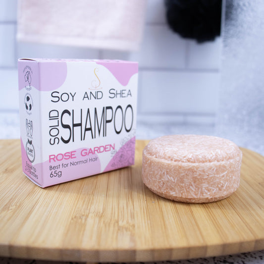 A white box with rose abstract pattern and the text SOLID SHAMPOO ROSE GARDEN stands upright on a wooden tray with a blurred bathroom background. Next to the box sits a round bar of solid shampoo. The bar is light pink with small white lines throughout.