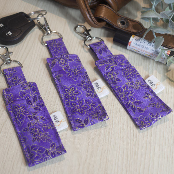 A selection of three clip balms showing variations in fabric design, lie on a wooden side with the middle Clip Balm attached to a car key. A lip balm lays near-by along with some greenery. The Clip balms are a rich puple colour featuring a floral design , as shade darker and outlined in gold