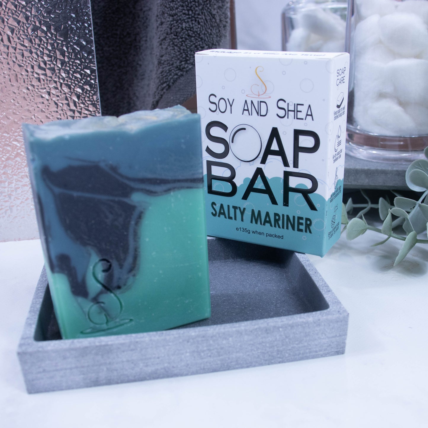 A close up look of the soap which features three layers of colour; a sea green base, black centre and teal at the top, which have been swirled together to look like a rolling wave. The soap is rectangular in shape and is taller than wide. It is sitting on a stone soap dish and behind it is the box the soap came in. The lower portion of the box has a bubble border coloured in sea green whereas the top is white with small grey bubbles. The box advises it is soap and the scent.