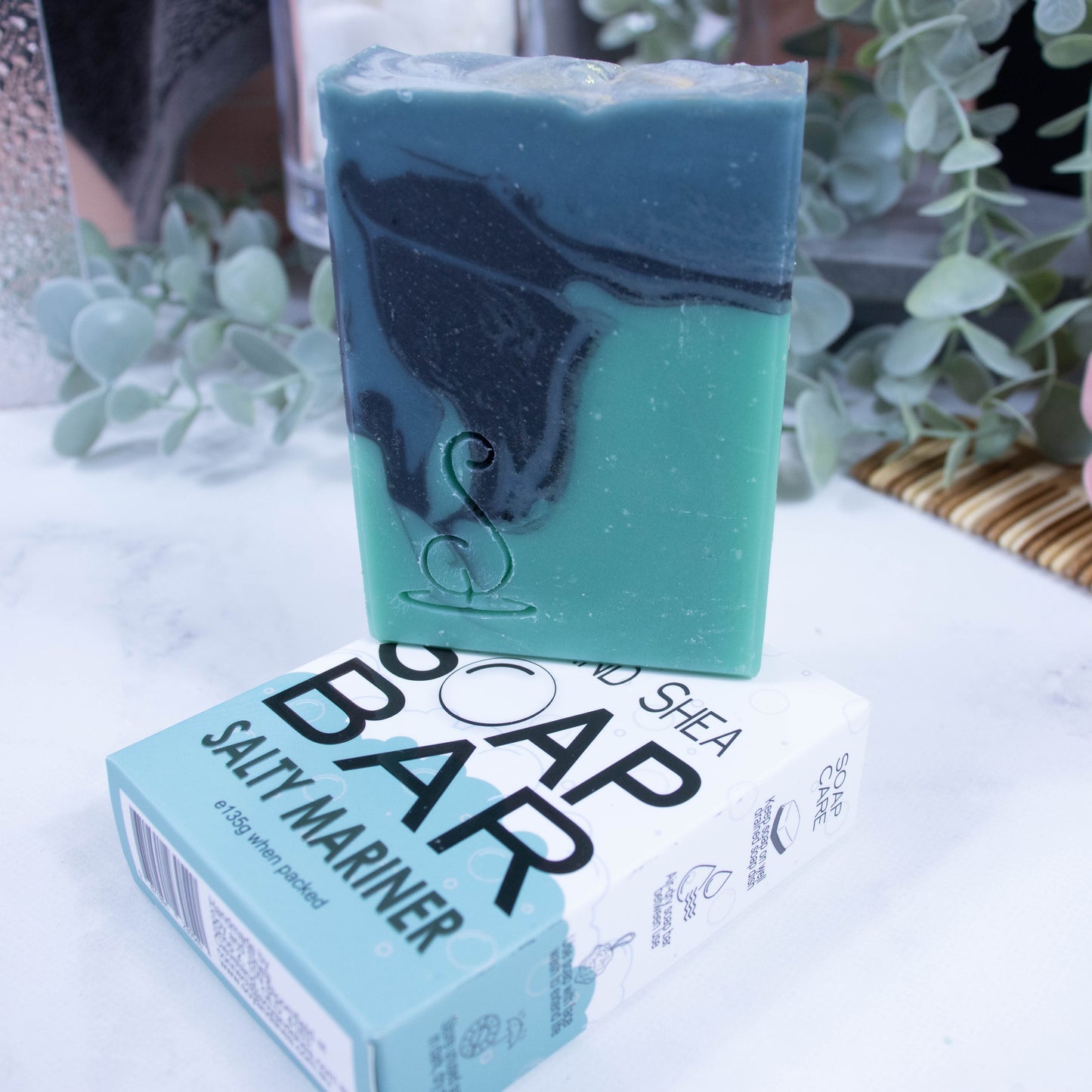 A tall rectangular soap bar is sitting on top of it's box.  The soap has three layers of colour; a sea green base, black centre and teal at the top, which have been swirled together to look like a rolling wave.  The soap box is laying flat and is white and sea green in colour
