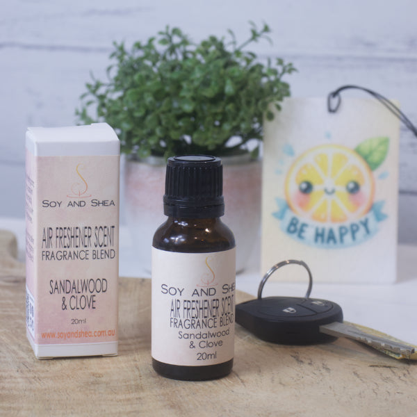 A small amber bottle with black lid labelled AIR FRESHENER SCENT stands on a wooden surface next to the box packaging. In the background is a car key, air freshener and greenery.