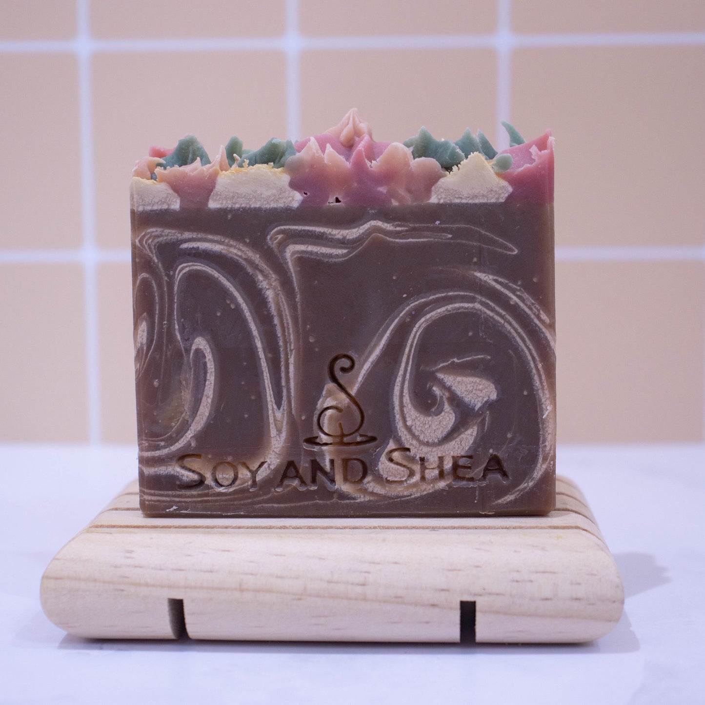 A bar of soap stands on a wooden soap dish on white surface with a faded peach tile background. The Soap has a main colour of chocolate brown with cream swirls. The top has piped soap in cream, pink and green