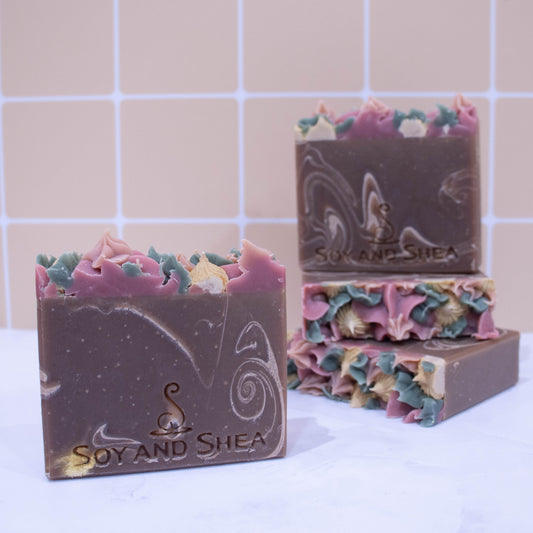 A collection of soaps sit on a white marble surface against a pale peach tile background. In the background, two soaps lay flat but stacked upon each other revealing a top of piped soap in shades of pink and cream. A third soap stands on stop of this stack with a fourth soap in the foreground. The soaps have a brown basee colour with cream swirls. Each soap shows a variation in swirl design