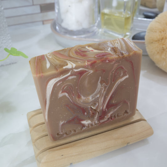 A bar of soap stands on a wooden soap dish with a blurred bathroom background. The soap is a light brown with swirls of dark brown, dark red and white swirls. The focus point is the top of the soap which is flat with coloured swirls.