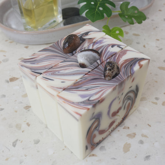 A selection of three soap stand on a row on a marble benchtop. The soaps are white with swirls of colour on top in shades of brown, black and purple. Each soap has a sardonyx stone in the top centre of each soap, each with a different look.