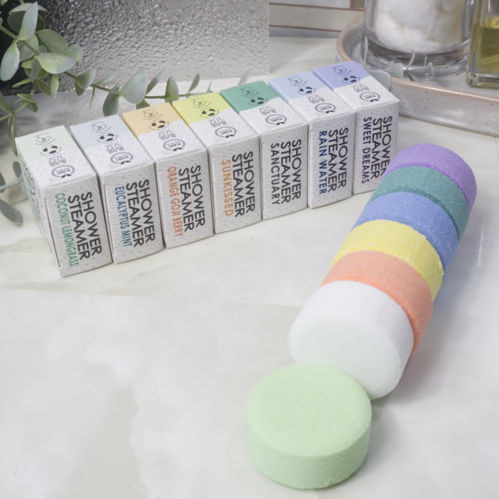 Seven small rectangular boxes of shower steamers are lined up behind a stack of round shower steamer discs in pastel colors, with one pale green pad placed in front on a light marble-like surface.