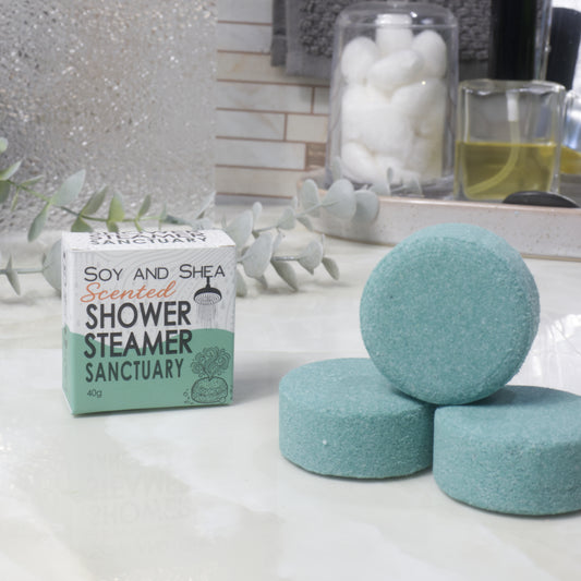 Three round dark green shower steamer discs displayed on a white marble surface next to their branded cardboard box, with decorative eucalyptus leaves and bathroom accessories blurred in the background.
