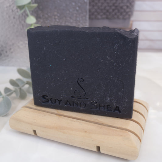 A solid black soap bar sits on a wooden soap dish with a shower screen and towel in the background