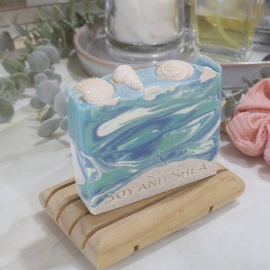 A soap bar sits on a wooden soap rack on a bathroom counter with a jar of cotton balls and perfume bottle in the background. The soap has a beige base layer topped with a marbled blue and white top layer made to look like water. The top is decorated with beige seashells
