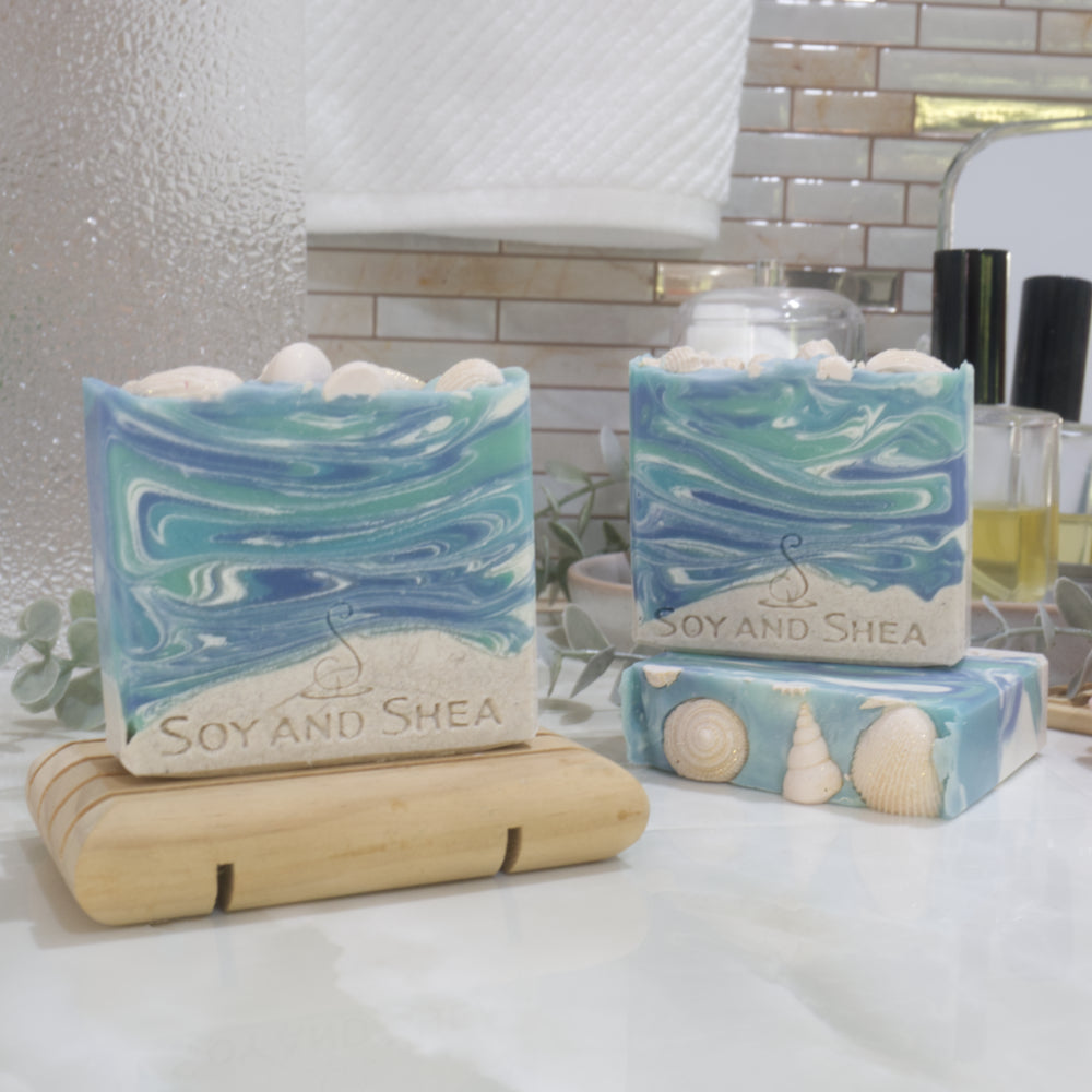 Three soap bars are displayed on a bathroom countertop. One soap lies flat on the marble surface showing beige soap seashell decorations on the top. Another soap stands on top of this bar with another to the left standing on a soap dish. The soaps have a beige base layer topped with a marbled blue and white top layer made to look like water. The top is decorated with beige seashells