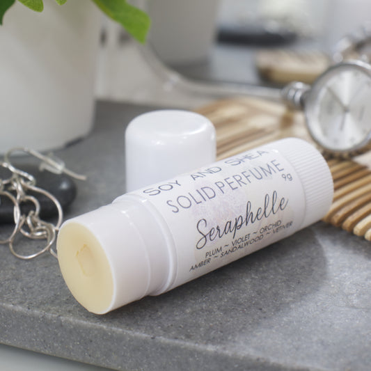 A white wind-up tube lays flat on a stone tray surrounded by accessories like earrings, watch and comb. The lid has been removed and placed behind the tube revealing a beige balm.