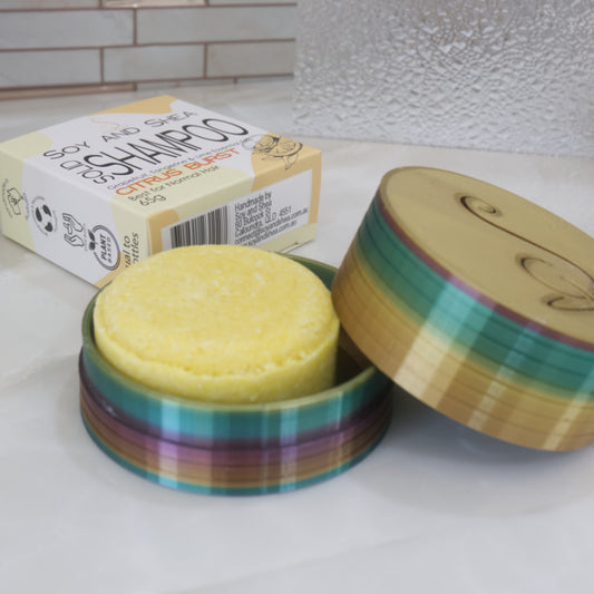 A yellow solid shampoo sits in a colourful round container with the cardboard packaging sitting behind.