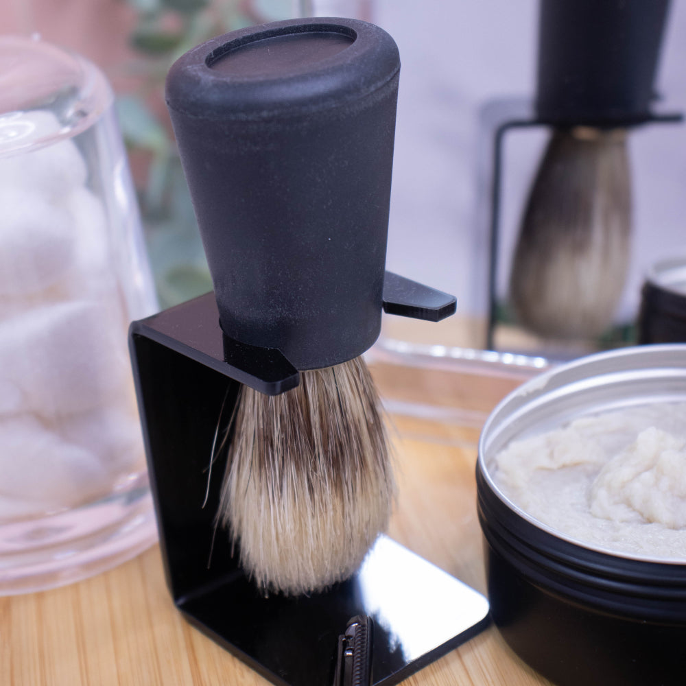 A black handled shaving brush is hanging on a plastic stand next to a open pot of shaving soap