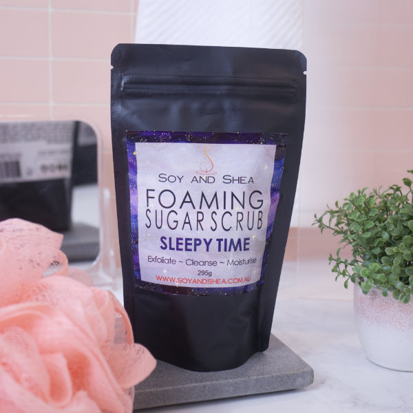 A black zip lock pouch stands upright on a stone tray in front of blurred bathroom background with peach tiles. The pouch, which stands between a potted plant and a loofah, has a label that reads FOAMING SUGAR SCRUB SLEEPY TIME and features a dark galaxy background with golden speckles for stars
