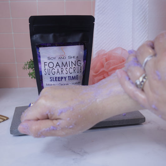 A hand applies the a purple sugar scrub to the back of the other hand and arm. In the background sits a black pouch of the scrub with a label depicting a night sky against a tile background with white towel