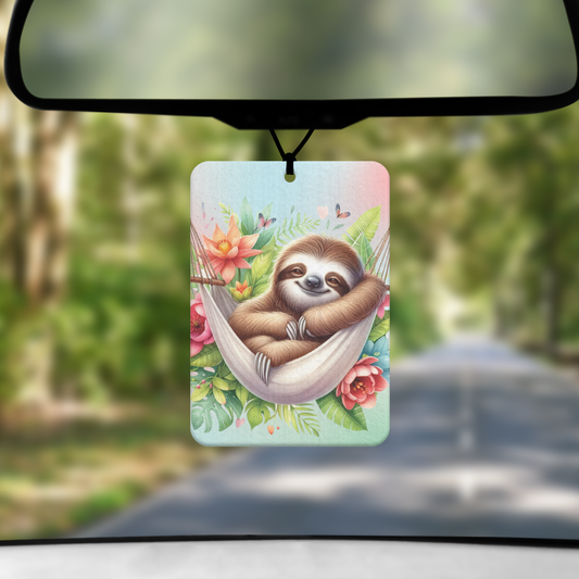A mock up image showing a view through a car windscreen with a long tree lined road. From the rearview mirror hands an air freshener with an image of a sloth in a hammock surrounded by tropical plants
