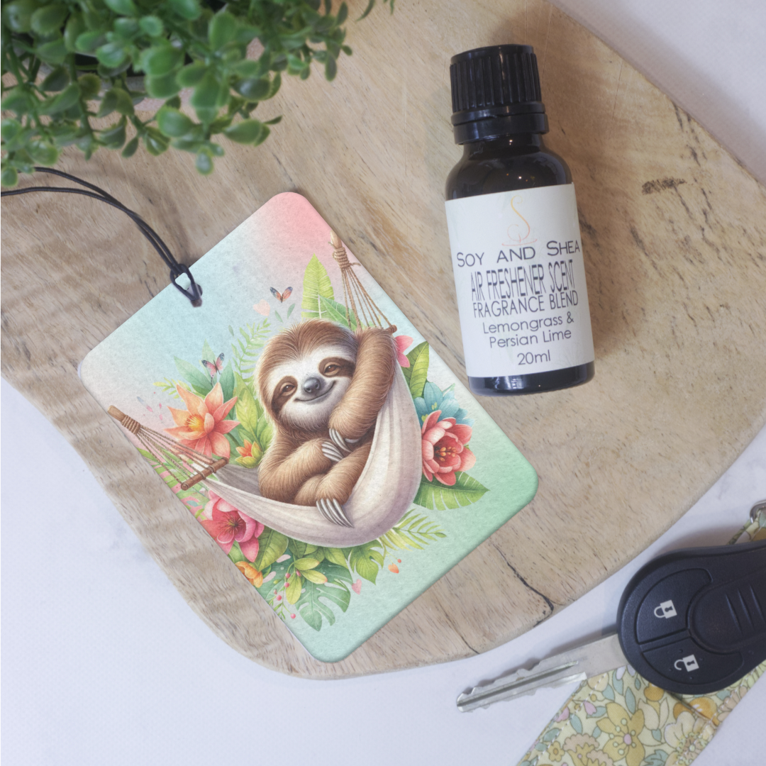 A Car Air Freshener lies on a wooden board next to a bottle of Air Freshen scent and a set of keys. The Air freshener shows a cartoon image of a sloth in a hammock surrounded by flowers