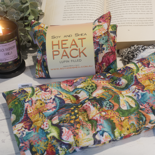 Two Small Heat packs are displayed on a wooden bench, with one laying out flat while the other sits behind with a belly band label reading HEAT PACK LUPIN FIlled. An open book, burning candle and greenery can be seen in the background evoking a cosy feel. The Heat packs feature a variety of watercolour dinosaurs tightly packed in and row after row. Jungle vines can be seen between some dinosaurs.