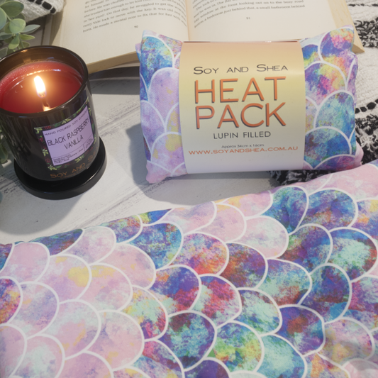 Two Small Heat packs are displayed on a wooden bench, with one laying out flat while the other sits behind with a belly band label reading HEAT PACK LUPIN FIlled. An open book, burning candle and greenery can be seen in the background evoking a cosy feel. The Heat packs have a mermaid tail design in shades of pink and purple.