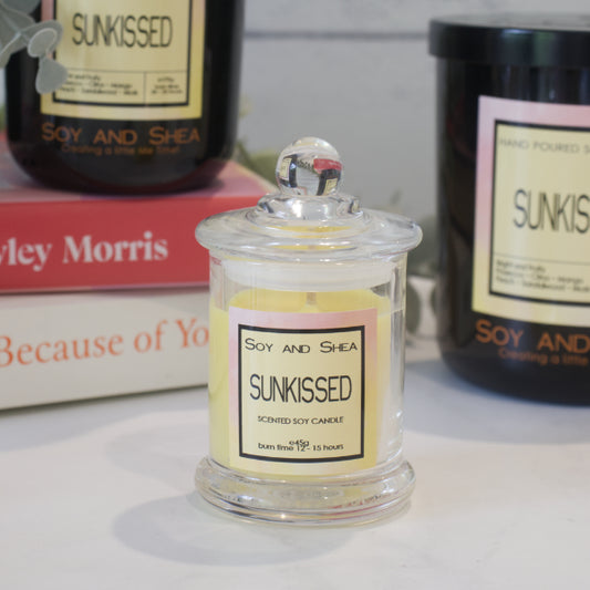 A small, clear glass candle jar with yellow wax sits in the foreground with a label reading SUNKISSED. Behind is a stack of books with a black candle resting on top with another large one to the right.