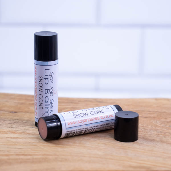 Two black tubes of lip balm are displayed on a wooden surface with blurred tiled background. One tube stands on its end while the other lays flat in front with the cap removed revealing a light red balm. The label has a blue background.