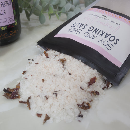 A black pouch of Soy and Shea Soaking Salts with a pink label spilling salt with rose petals across a marble bench