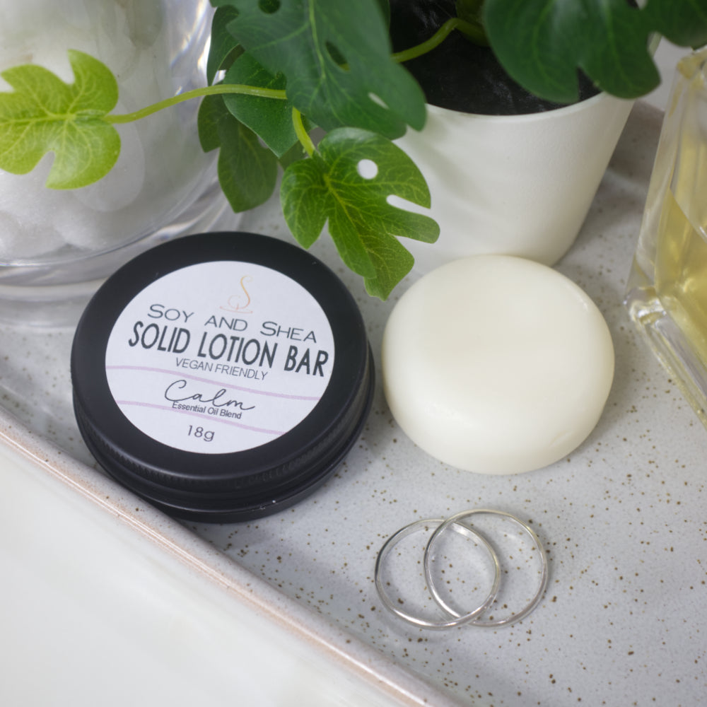 A small black round tin sits on a stone tray with a label reading SOLID LOTION BAR. Next to it is a solid white bar of lotion. The items are surrounded by jewellery, a potted pland and a bottle