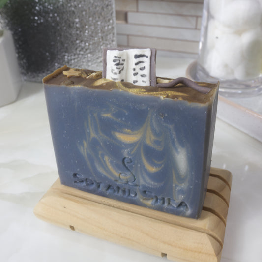 A soap bar stands on a wooden soap dish on a bathroom counter. The soap is dark blue with gold and white streaks of colour throughout. The top of the soap features embellishments made of soap including an open book standing up on the soap along with a wand and gold stars.