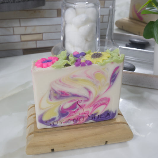 A rectangular bar of soaps stands on a wooden soap dish on a bathroom countertop. The soap has a cream base colour with swirls in neon pink, dark purple and bright yellow. The top features a collection of brightly coloured flower embellishments including a sunflower