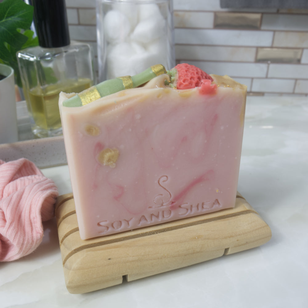 A bar of soap stands upon a wooden soap dish on a bathroom countertop. The soap is pale pink with dark pink and gold swirls. The top of the soap features soap embeds of a green champagne bottle and a red strawberry