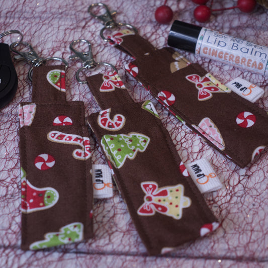 A selection of three clip balms showing variations in fabric design, lie on a white surface with a red mesh with one Clip Balm attached to a car key with a lobster clip. A lip balm stands near-by along with some Red berries. The Clip balms are brown with various christmas elements including candy can\es, bells, stockings and Christmas trees.