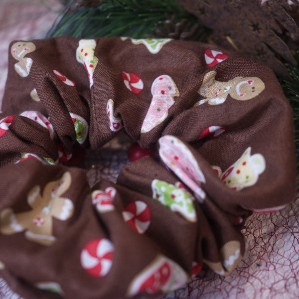A close up image of the fabric on the Sugar and Spice Scrunchie. It has a rich brown background covered in images of Christmas themed sugar cookies like trees and gingerbread men along with peppermint swirl candies