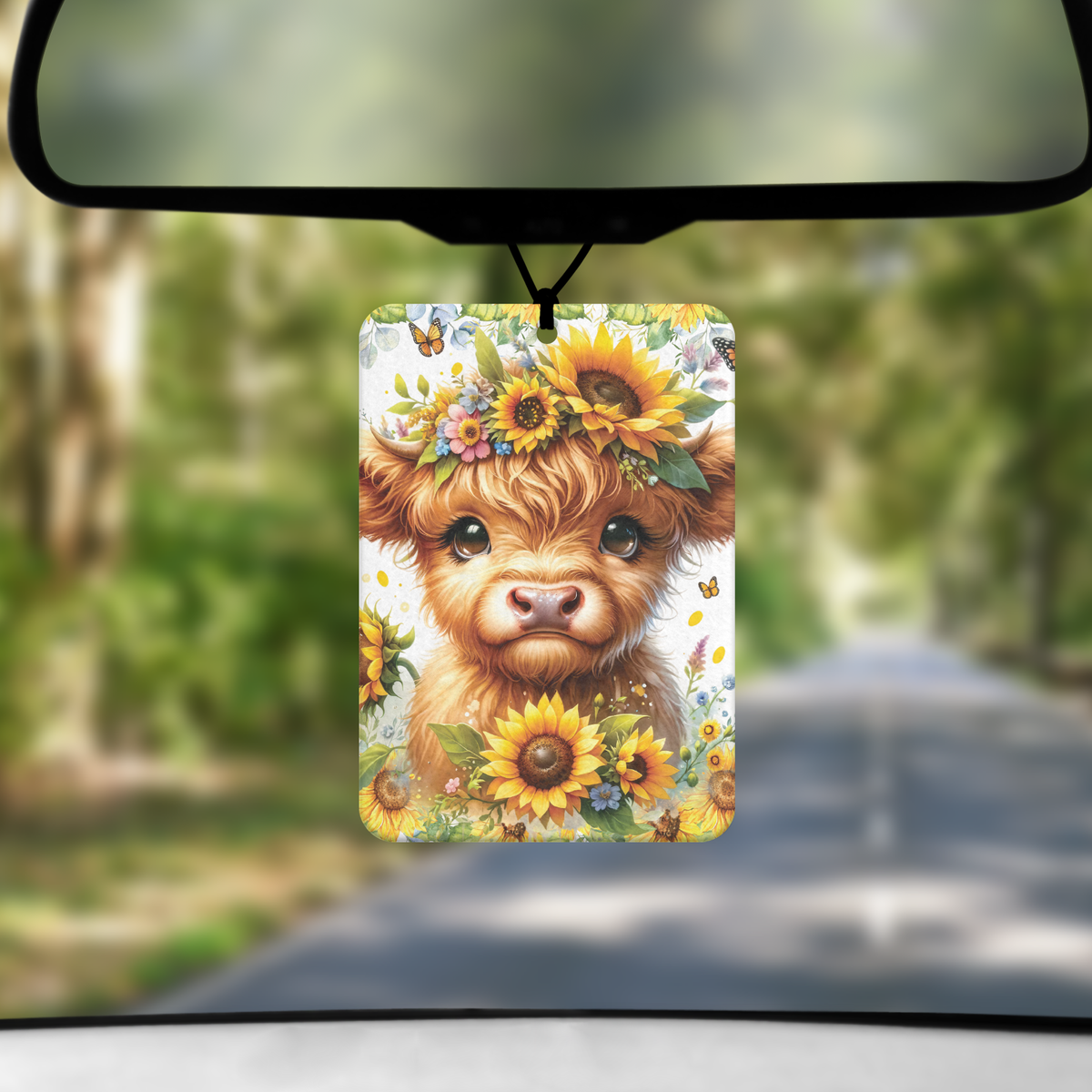 A mock up image showing a view through a car windscreen with a long tree lined road. From the rearview mirror hangs an air freshener with an image of a baby highland cow's head surrounded by sunflowers and butterflies