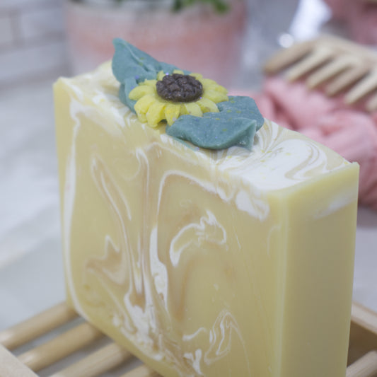 A close shot of a yellow bar of soap with white swirls showcasing a sunflower decoration on the top of the soap.