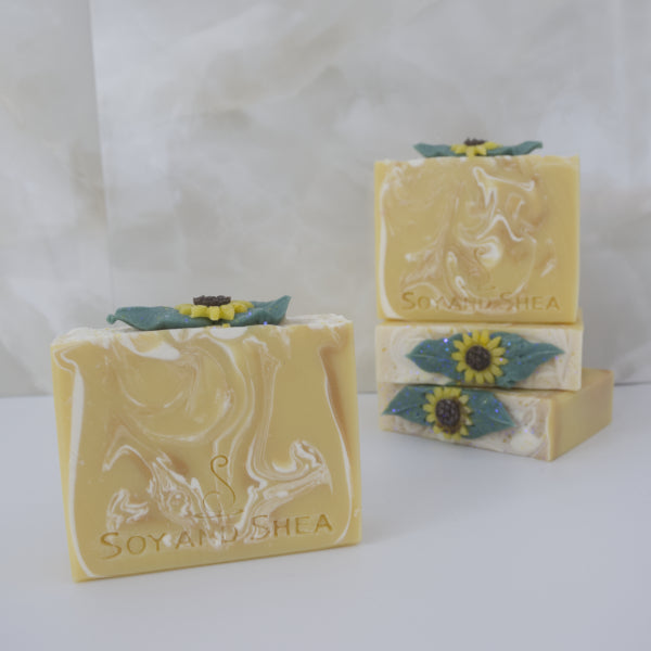 A selection of soaps sit on a marble tile background. Two soaps lie on their sides stacked on each other showing each soap with a decorative Sunflower embellishment. On top of these is a soap standing up showing a bar that is yellow with white swirls. To the front of these is another soap bar with the same colours but different pattern