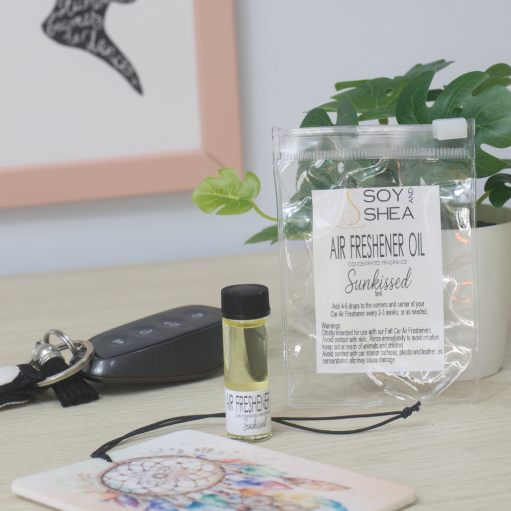 A small clear bottle with black cap of Soy's Shea Air Freshener Oil next to a clear resealable pouch, displayed on a light wood tabletop with a car key, a felt air freshener, and a potted plant in the background.