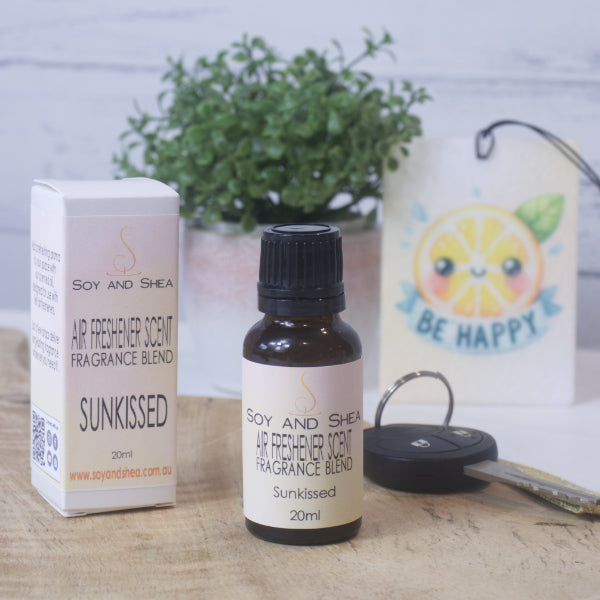 A small amber bottle with black lid labelled AIR FRESHENER SCENT stands on a wooden surface next to the box packaging. In the background is a car key, air freshener and greenery.