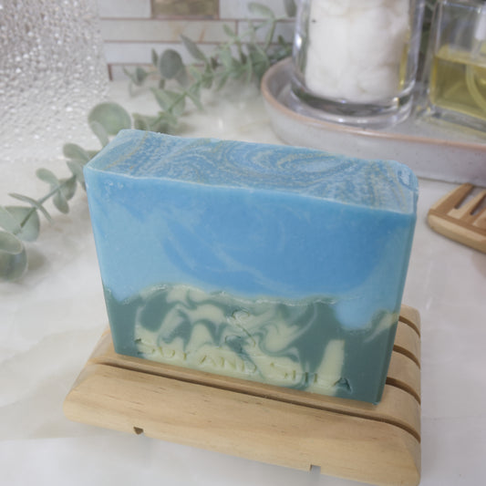 A bar of soap stands on a wooden soap rack on a bathroom counter top with a blurred background of greenery and bottles. The soap has two layers with the bottom half having a swirl of light and dark green lookin glike a mountain side. The top has a swirl of bright and light blue. The top of the soap is flat with gold swirls through the blue