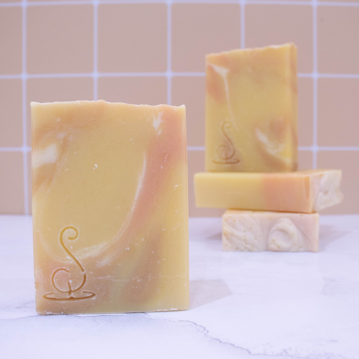 Four rectangular bars of soap sit on a marble bench each showing various design patterns from the same batch of soap. The soaps are a sunshine yellow colour and have streaks of dark gold and white.  All soaps have a similar pattern but all have slight differences